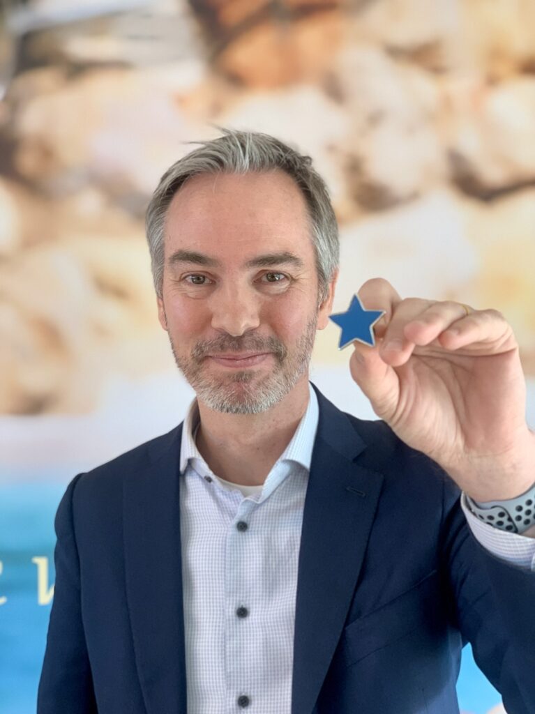 A man in a suit smiles and holds a small blue star towards the camera, with a blurred beach-themed background behind him.