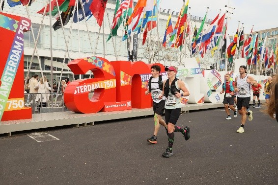 Runners participate in a race in front of the large red and white I amsterdam sign, surrounded by numerous international flags outside a modern building.