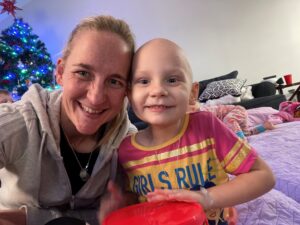 A smiling woman and a young child with no hair pose for a selfie indoors, capturing a heart-warming moment of Camilla's WishMaker Story, with a decorated Christmas tree and cosy blankets in the background.