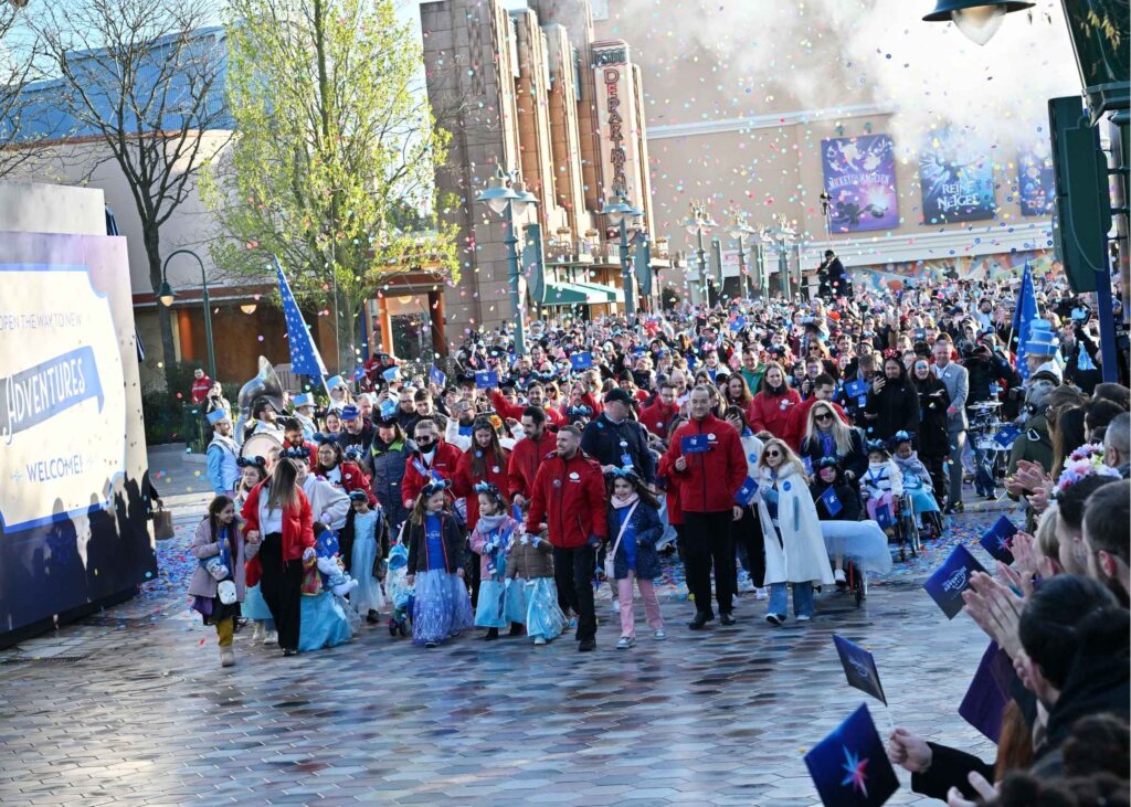A crowd walks down a decorated street with people waving flags and wearing fancy dress, amid confetti and celebration, as onlookers clap and cheer in an outdoor festive procession.