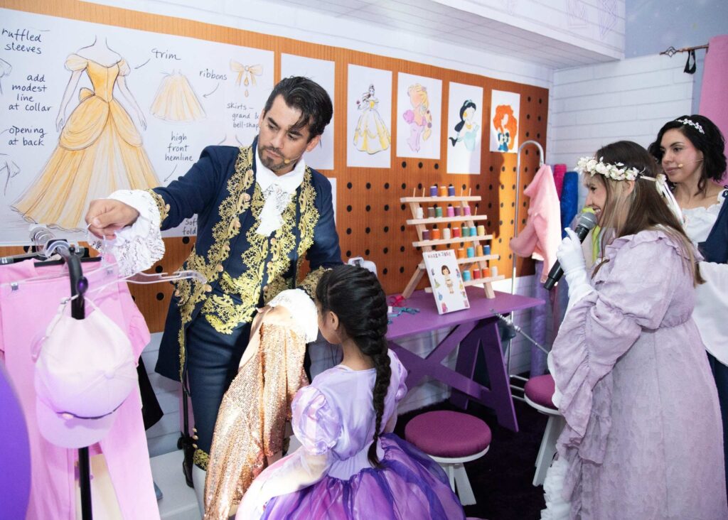 A man in a royal costume helps a young girl in a purple dress choose fabric, while two women in gowns look on, in a room decorated with dress sketches and sewing supplies.