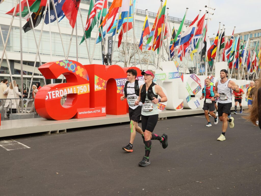 Runners in athletic kit pass by a large “I amsterdam” sign, with dozens of colourful international flags in the background, during a marathon event inspired by Camilla's WishMaker Story.