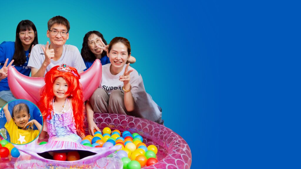 A group of smiling adults and children pose together; a girl in a red wig and mermaid costume sits in a small ball pit, surrounded by colourful balls, with others making peace signs and smiling at the camera.