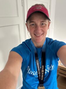 A person wearing a pink cap, blue Make-A-Wish shirt, and a medal on a black ribbon smiles for a selfie indoors, capturing a moment from Camilla's WishMaker Story. A white door and part of a wooden table are visible in the background.