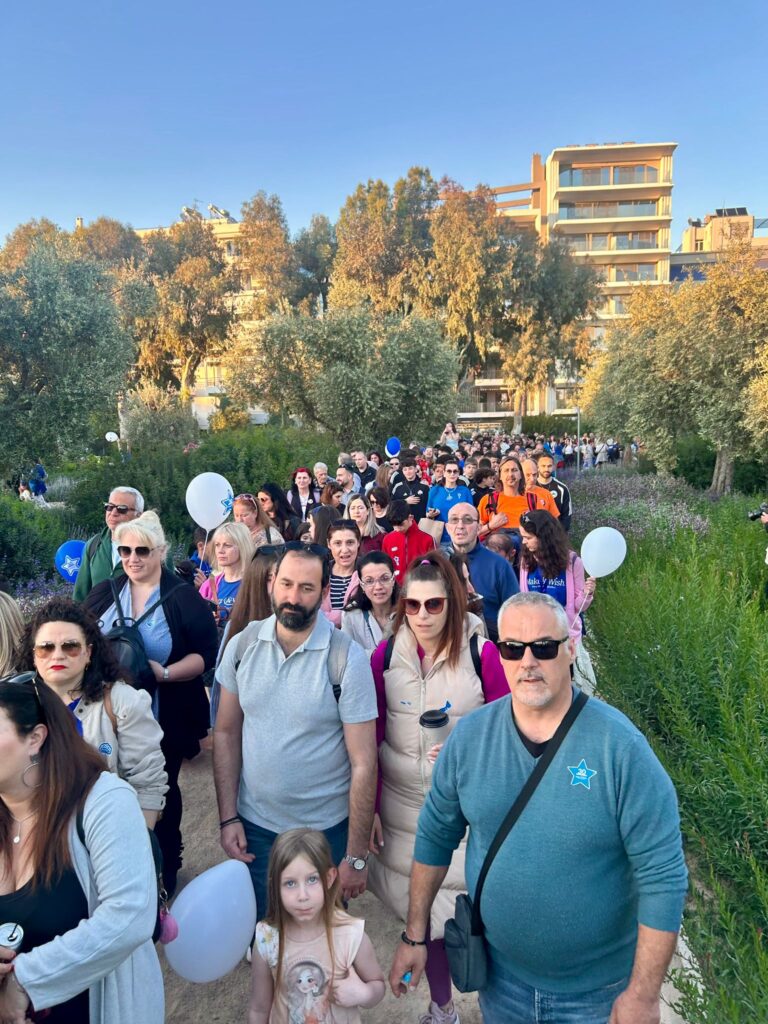 A large group of people, including adults and children, walk along a path surrounded by greenery. Some hold white balloons. The sky is clear and buildings and trees are visible in the background.