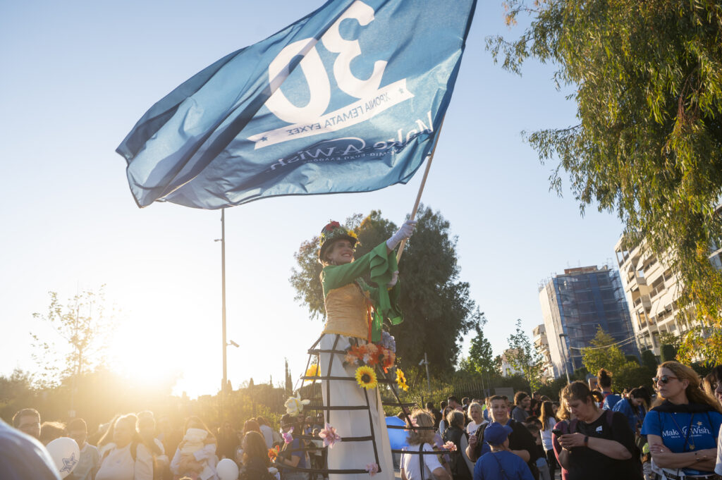 A woman on stilts in a flower crown and a costume with sunflowers waves a large blue flag marked 30 above a crowd at an outdoor event during sunset. Trees and modern buildings are visible in the background.