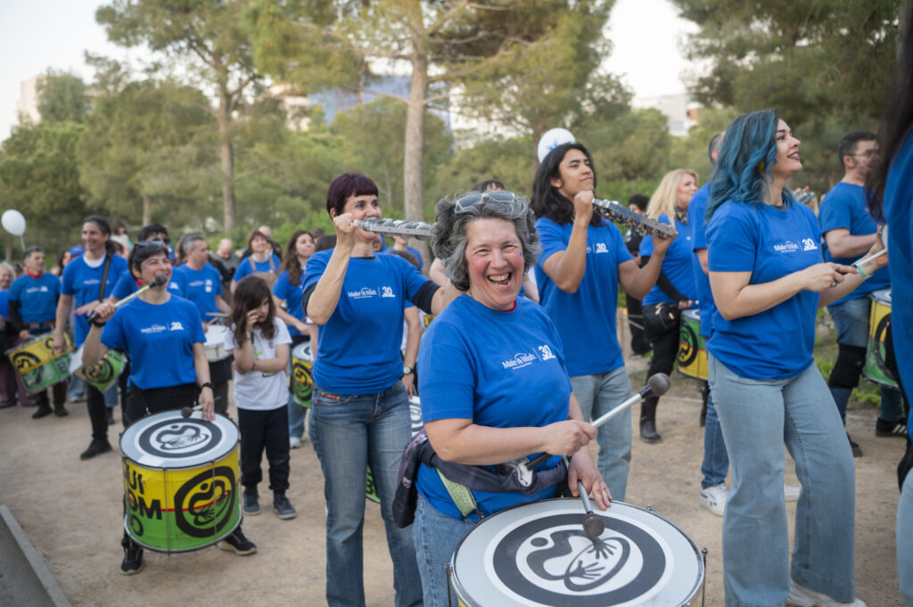 A group of people in matching blue shirts play drums and flutes outdoors, smiling and enjoying themselves among trees in a park setting.
