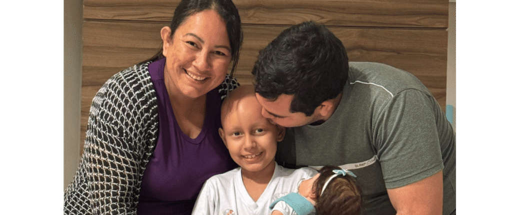 A smiling woman and man embrace a young child with a bald head; the man, wearing a Make-A-Wish shirt, kisses the child’s head while holding a baby. They are sitting together in a cosy, indoor setting during World Wish Month.