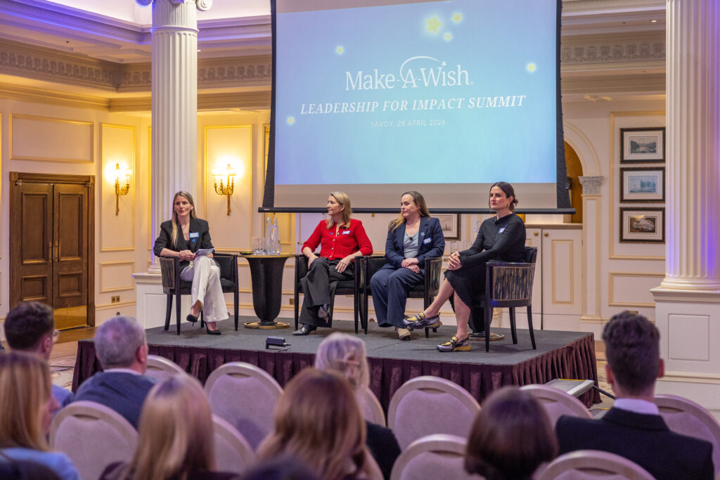 Four women sit on stage at the Leadership for Impact Summit in front of an audience. Behind them, a large screen displays the event name and World Wish Day date in an elegant, well-lit room.