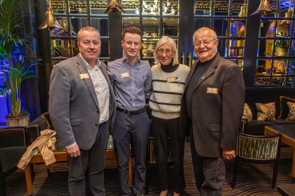 Four people stand closely together, smiling for the camera at the Leadership for Impact Summit in a stylish, warmly lit lounge with black and gold decor and indoor plants. Each person wears a name badge, a blue ribbon pin, and celebrates World Wish Day.