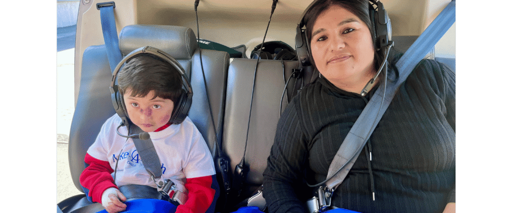 A child and an adult wearing headsets and seatbelts sit together in the back seat of a vehicle, possibly a helicopter, during a Make-A-Wish experience for World Wish Month. The child wears a white shirt with red sleeves; the adult smiles slightly in black.