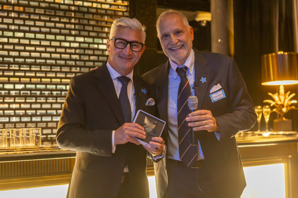Two men in suits stand smiling indoors at the Leadership for Impact Summit; one holds a glass award, and the other a microphone. The background features a wall of plaques and warm, ambient lighting, celebrating World Wish Day.