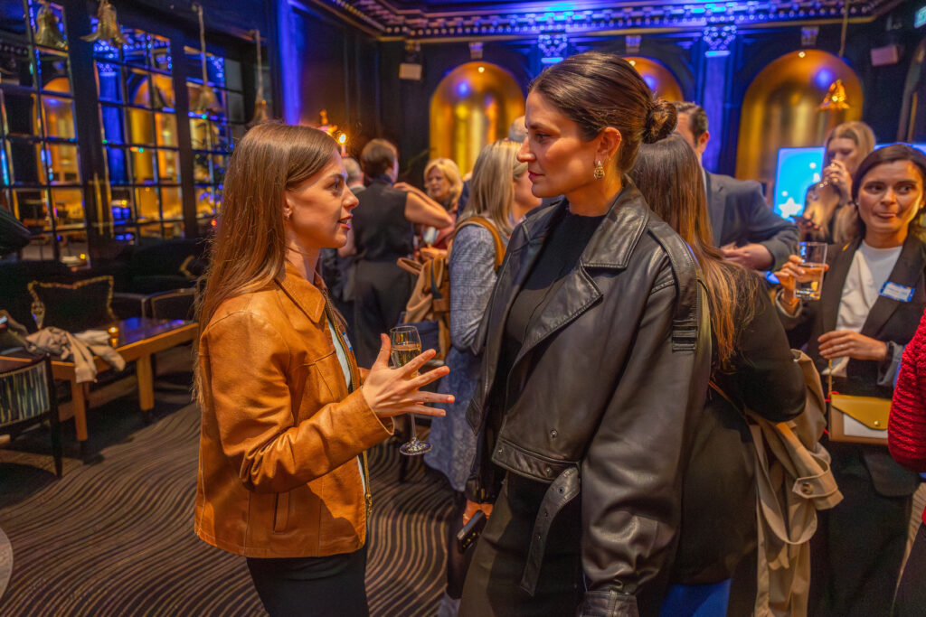 Zwei Frauen unterhalten sich im Innenbereich des Leadership for Impact Summit, jede mit einem Getränk in der Hand. Im Hintergrund mischen sich weitere Gäste vor dunklen Wänden, warmem Licht und in einer lebhaften, eleganten Atmosphäre.
