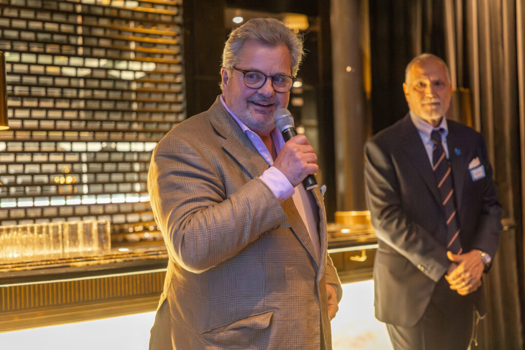 At the Leadership for Impact Summit, a man in a beige suit and glasses speaks into a microphone at an indoor event, whilst another man in a dark suit stands in the background near a lit bar area on World Wish Day.