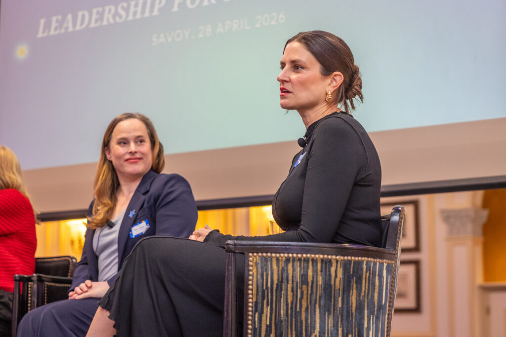 Two women in professional attire sit on stage during the Leadership for Impact Summit. One, in a black dress, speaks as the other, in a navy suit, listens. The projection behind them highlights World Wish Day and event details for 28 April 2026.