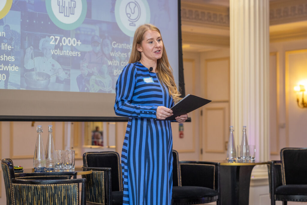 A woman in a blue and black striped dress speaks on stage at the Leadership for Impact Summit, holding a folder. Behind her, a presentation screen displays graphics and text. Four water bottles and glasses sit on a table in the elegant conference room.