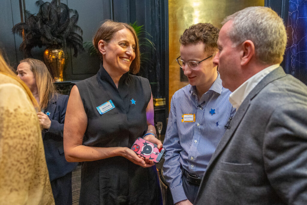 Beim Leadership for Impact Summit unterhält sich eine lächelnde Frau, die einen kleinen Roboter hält, mit zwei Männern in einem eleganten Veranstaltungsraum. Alle drei tragen Namensschilder; die anderen Teilnehmer sind vor dem gold-dunklen Dekor gut zu erkennen.