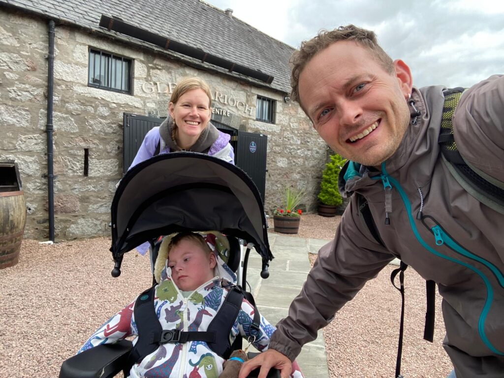 A man and woman smile for a selfie outdoors, standing behind a child in a pushchair. The child, wearing a colourful jacket and dreaming of Ebranaz's wish to be a fairy, looks down. They are in front of a stone building with a sign.