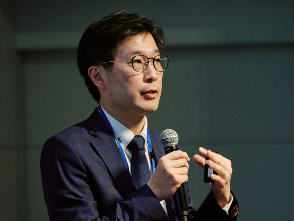 A man wearing glasses and a dark suit speaks into a microphone whilst gesturing with his other hand during a presentation or conference.