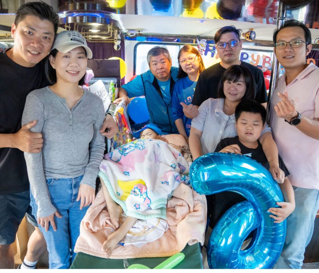 A group of people pose together at a birthday celebration for World Wish Month, with one person in bed covered by a cartoon blanket and another holding a large blue number 6 balloon. Make-A-Wish decorations and balloons are in the background.