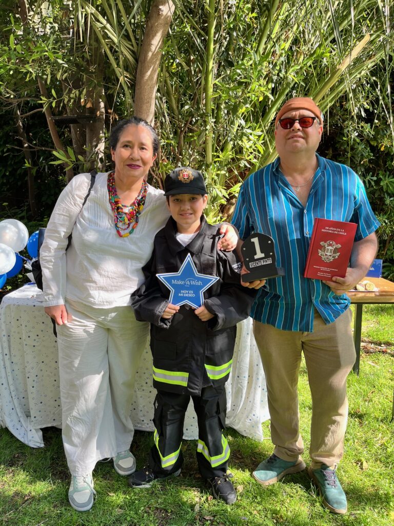 Three people stand outdoors on grass during World Wish Month: a woman in white, a child dressed as a firefighter holding a star-shaped award, and a man in sunglasses with a plaque and red book. Balloons and trees hint at the Make-A-Wish celebration.