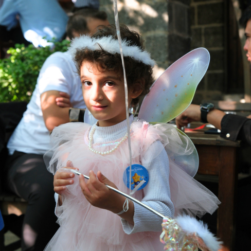A young child dressed as a fairy, wearing pink tulle, wings, a headband, and holding a wand, stands outdoors among people, with sunlight shining on their face.