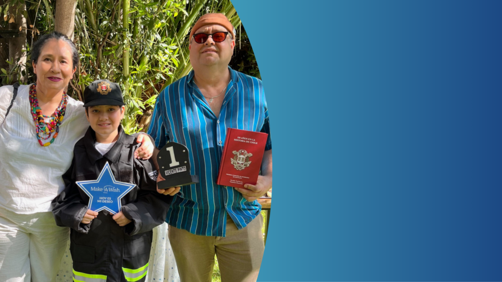 A smiling family stands outdoors for World Wish Month. A woman in white is on the left, a child in a firefighter costume holds a blue star award, and the man in stripes proudly displays his Make-A-Wish trophy and red folder. Right side has a blue gradient.