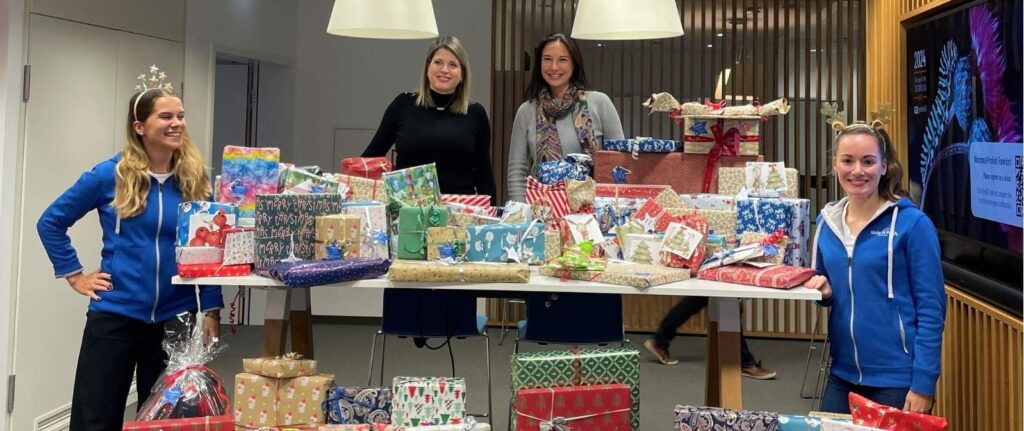 Four women stand smiling behind a table covered with gifts in a festive office, celebrating Robert Half fundraising efforts for Make-A-Wish. Holiday-themed headbands add to the cheerful, celebratory atmosphere.