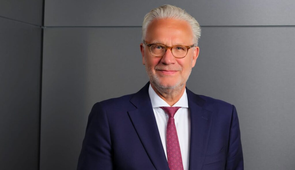 Wijnand Donkers, Chair of its Board of Directors, with grey hair and glasses, wears a navy blue suit, white shirt, and red patterned tie as he smiles in front of a plain grey background.