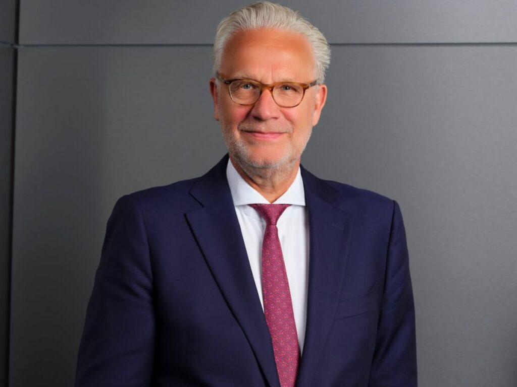 An older man with white hair and glasses, wearing a navy blue suit, white shirt, and red patterned tie, stands in front of a plain grey background, smiling at the camera.