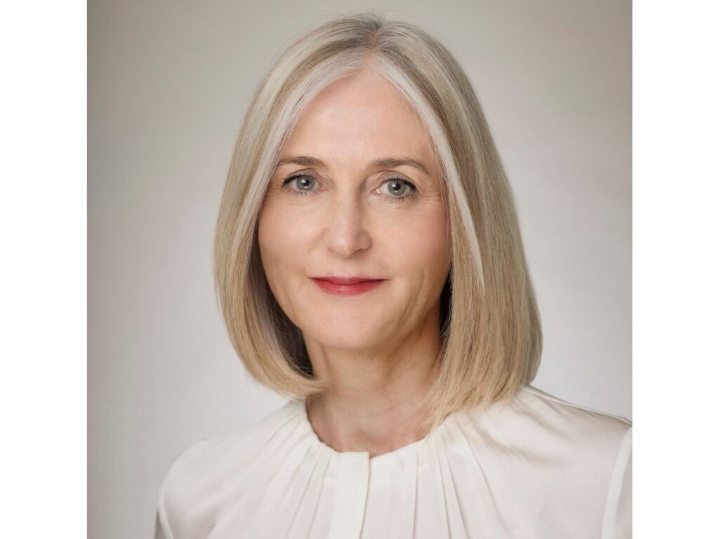 A woman with straight, shoulder-length grey-blonde hair and fair skin, wearing a cream-coloured blouse, smiles softly at the camera against a plain light background.