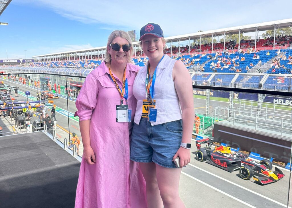 Two women pose and smile at a Formula 1 race track, with Red Bull F1 cars and team garages visible behind them. One wears a pink dress and sunglasses, the other wears a hat, sleeveless top, and denim shorts. Spectators sit in the stands.