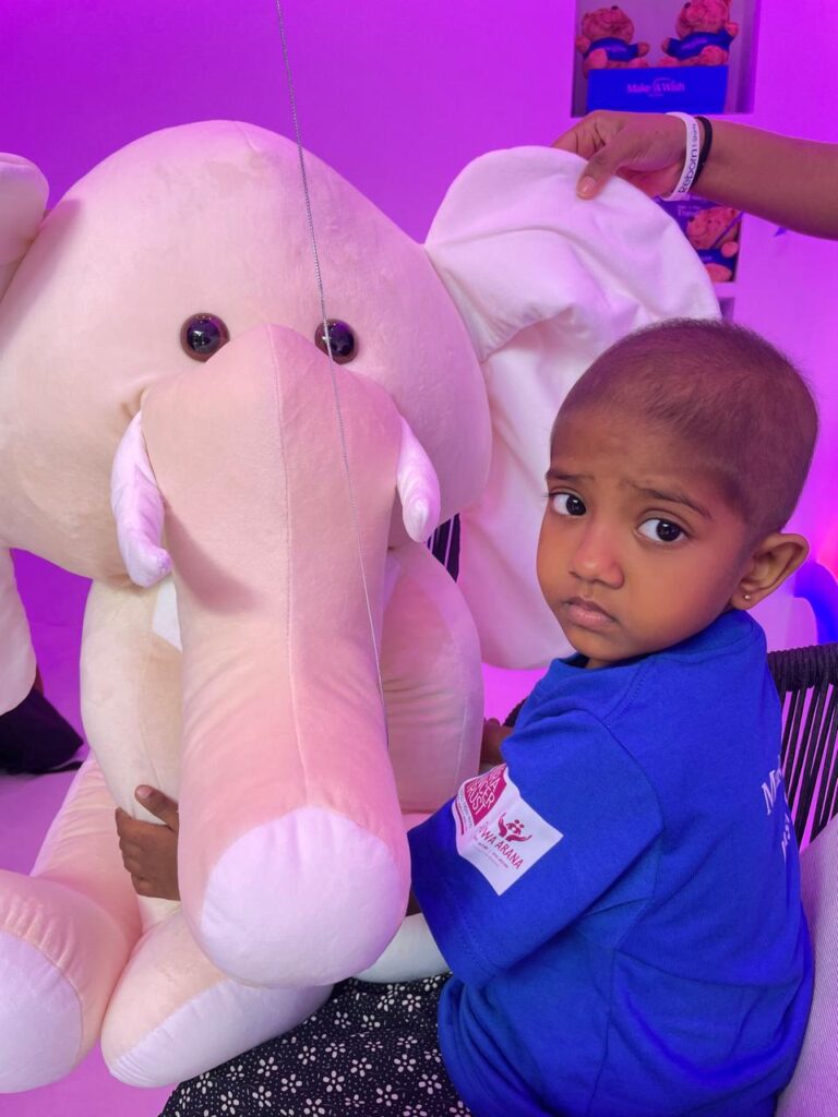 A young child in a blue shirt sits with a large, pale pink stuffed elephant, looking at the camera—perhaps dreaming of Luca’s wish to fly in a helicopter. An adult’s hand rests gently on the elephant. The background is lit with purple light.