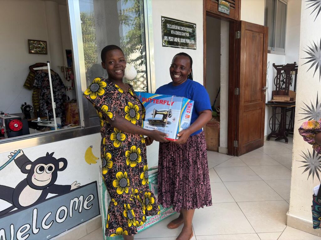 Two women smiling and holding a boxed sewing machine together inside a bright room. One wears a floral dress, the other a blue shirt and patterned skirt. A Welcome sign with a cartoon monkey is visible nearby.