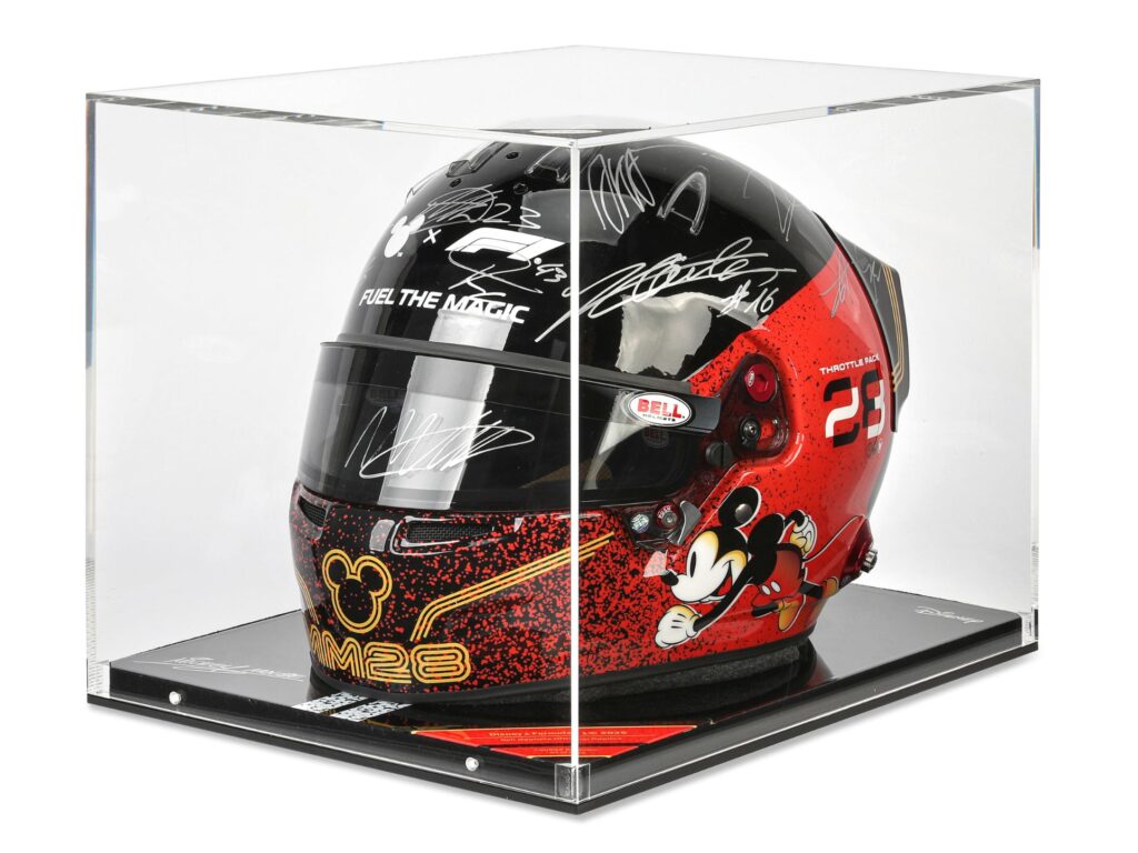 A black and red racing helmet with Mickey Mouse graphics and several signatures, displayed in a clear acrylic case on a black plinth.