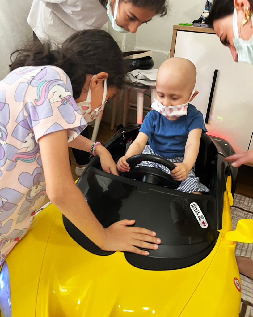 A young child wearing a mask sits in a yellow toy car, holding the steering wheel. Two women and another masked child stand nearby, gently supporting the child and car, as they listen to Binaya's wish to have a soft elephant toy.