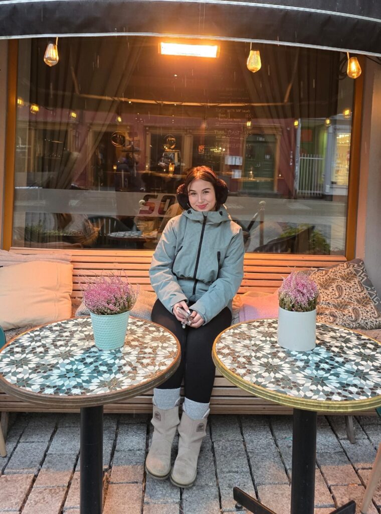 A woman in a grey jacket sits on a wooden bench between two round tables with potted flowers outside a cosy café. Warm lights hang above, and cushions rest beside her.