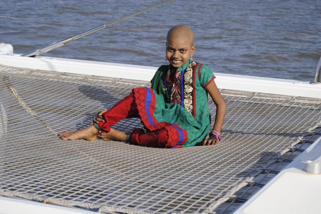A smiling child with a bald head wears colourful traditional clothes and sits cross-legged on a netted area of a boat, with water visible in the background, dreaming of Jerida’s wish to have a sewing machine fulfilled.