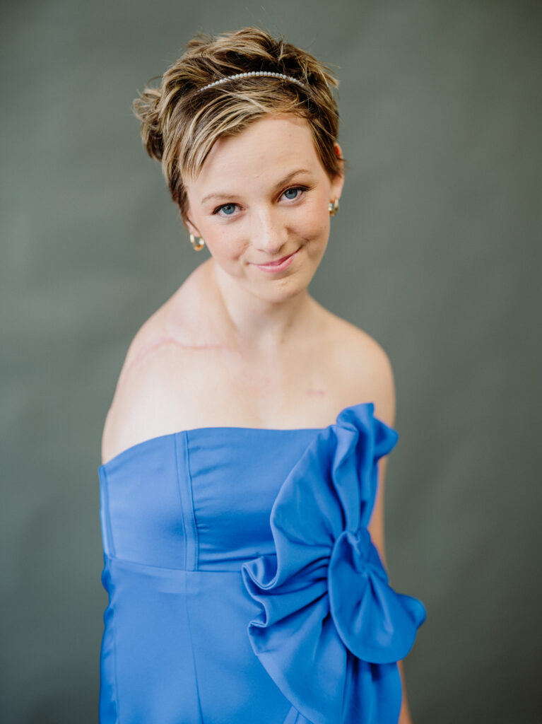 A young woman with short hair, wearing a strapless blue dress adorned with a large fabric flower, a hairband, and earrings, poses and smiles gently at the camera against a plain grey background, her joyful expression hinting at Rylee’s wish to have a shopping spree come true.