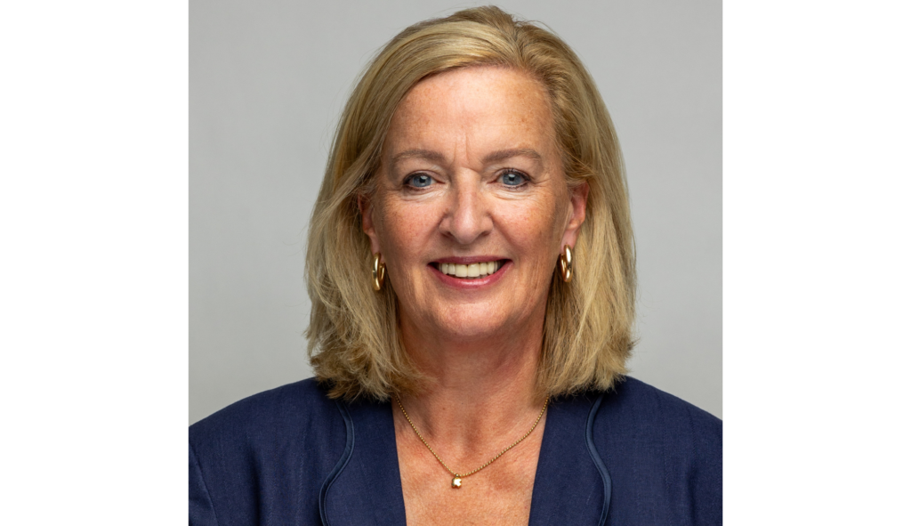 A smiling middle-aged woman with blonde hair wearing a navy blue blazer, gold earrings, and a gold necklace, against a plain grey background.