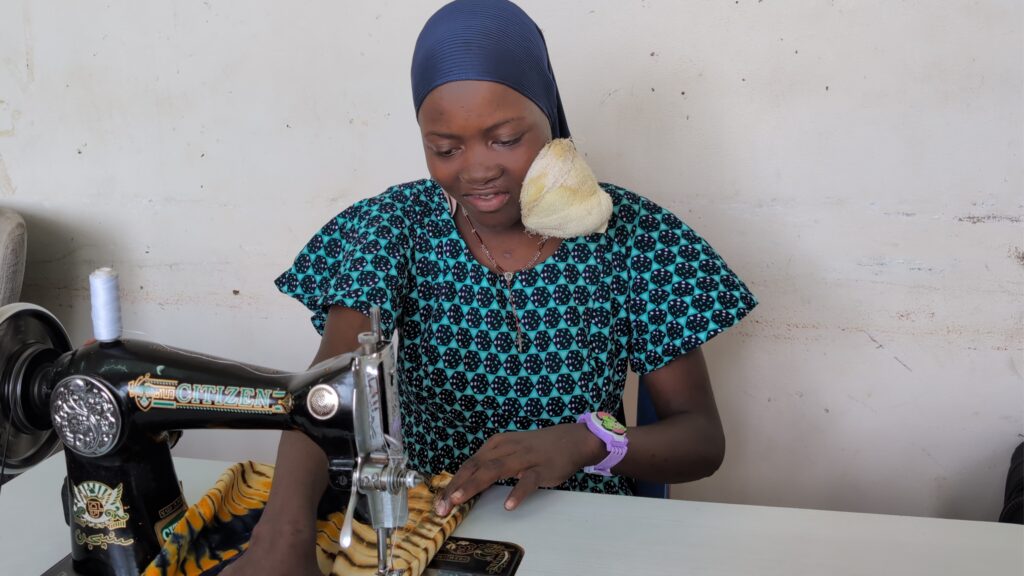 A woman in a patterned dress and headscarf, with a cloth on her shoulder, uses a sewing machine to stitch fabric. She is focused on her work at a table against a plain wall.