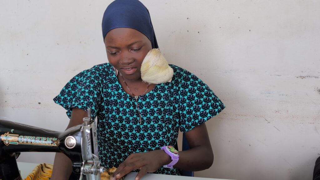 A woman wearing a patterned dress and headscarf operates a sewing machine. She has a large swelling on her jaw and is focused on sewing fabric, seated against a plain, light-coloured wall.