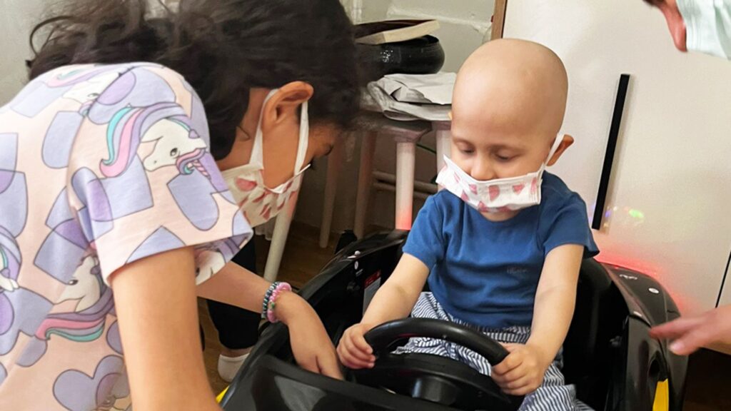 A young child wearing a mask and sitting in a toy car, inspired by Defne’s wish for a battery-powered car, holds the steering wheel whilst another masked child stands nearby, assisting or interacting with them. Both children appear to be indoors.