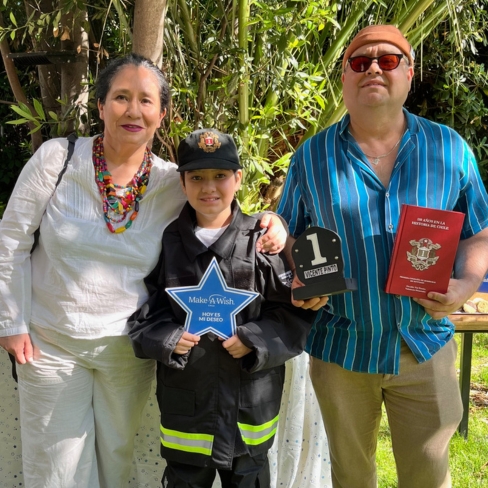 Three people pose outdoors. A child dressed as a firefighter holds a blue Make-A-Wish star. An adult on the left smiles, and an adult on the right holds a plaque and a red book. Greenery and sunlight are in the background.