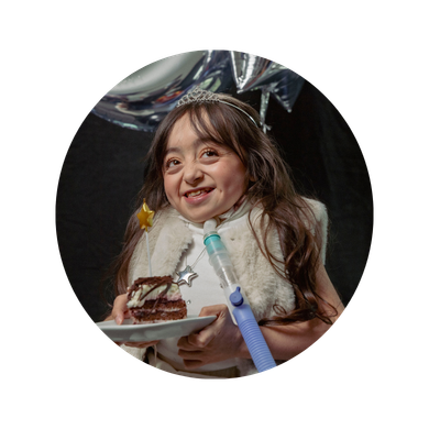 Young girl wearing a tiara and fur gilet, smiling and holding a plate with cake. She uses a ventilator and stands in front of balloons.