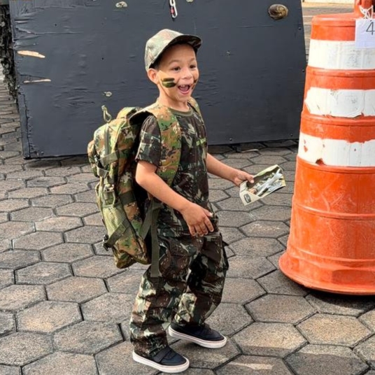 A young boy dressed in camouflage military attire with a rucksack and face paint smiles whilst holding paper, standing near an orange traffic barrel outdoors.