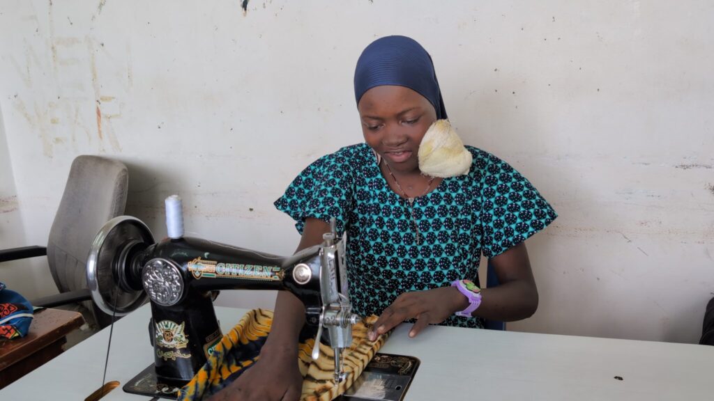 A woman wearing a blue patterned dress and headscarf sits at a sewing machine, sewing colourful fabric at a table in a simple room.