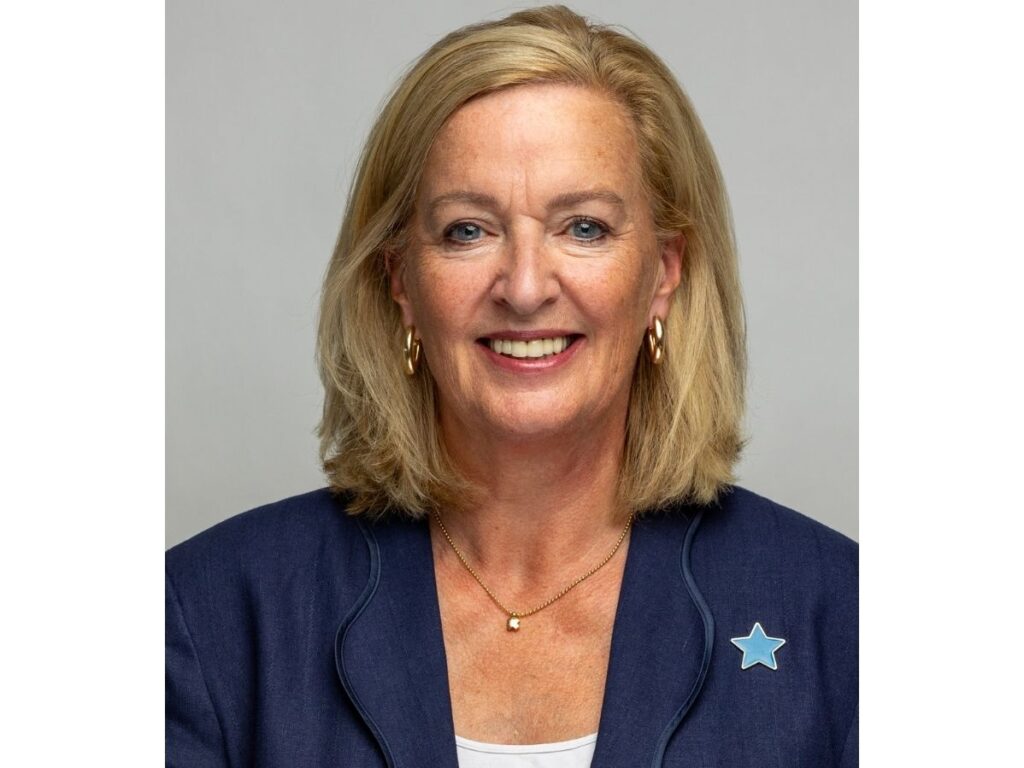 A smiling woman with shoulder-length blonde hair, wearing a navy blazer with a star pin and a gold necklace, poses against a plain light grey background.
