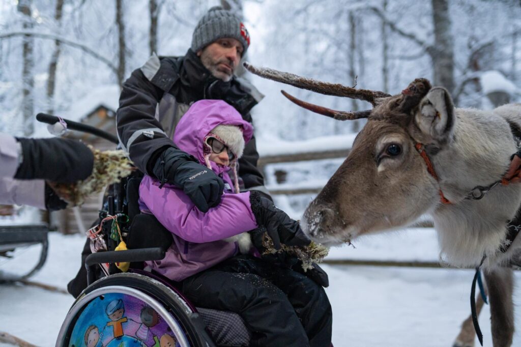 A man in a winter jacket and beanie assists Mía in her wheelchair as she feeds a reindeer in a snowy forest, fulfilling her wish to go ice skating.