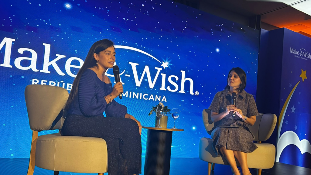 Two women sit in armchairs on a stage with a blue Make-A-Wish Dominican Republic backdrop, both speaking into microphones during the event as Make-A-Wish launches in the Dominican Republic.
